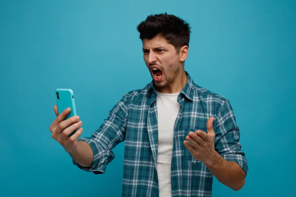 angry young man stretching mobile phone out looking it keeping hand air shouting isolated blue background